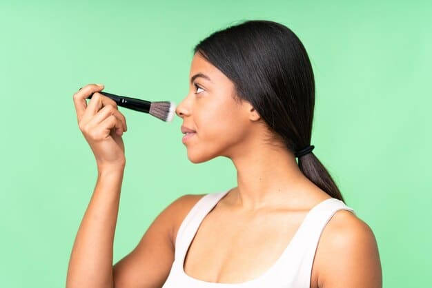 A woman applying highlighter to her cheekbone with a fan brush, demonstrating proper technique for a natural-looking glow.