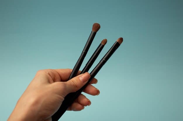 A close-up of a hand holding three different types of eyeshadow brushes: a fluffy blending brush, a flat shader brush, and a pencil brush. The image shows the texture and shape of the bristles, as well as the size and handle of each brush. The background is softly blurred to emphasize the brushes themselves