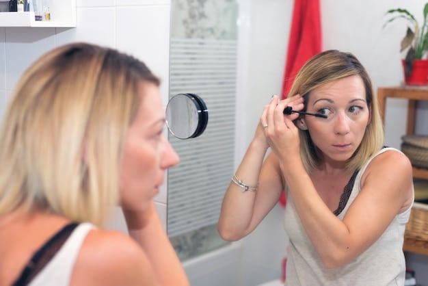 A woman applying waterproof mascara with a focused expression in a well-lit bathroom. Her makeup is natural, highlighting the mascara's effect on her lashes.