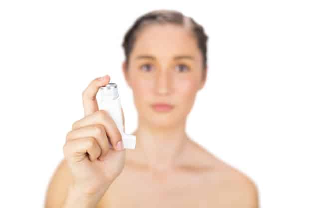 A woman with dry skin applying a hydrating setting spray. The spray bottle is close to her face, showing the fine mist. Her skin looks dewy and fresh. The background is a softly lit bathroom with skincare products on the counter.
